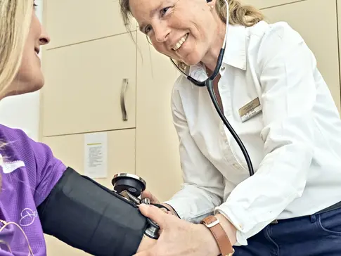 Ich sehe eine Ärztin mit weißer Bluse und dunkelblauer Hose, die eine blonde Patientin in lila T-Shirt mit Stethoskop und Blutdruckmanschette untersucht.