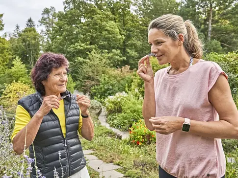 Hier sehen Sie zwei Frauen in einem Kräutergarten, die gerade Kräuter ernten und daran riechen.
