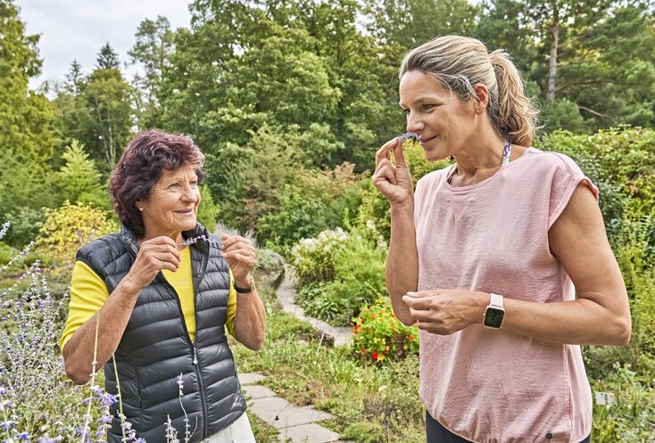 Hier sehen Sie zwei Frauen in einem Kräutergarten, die gerade Kräuter ernten und daran riechen.