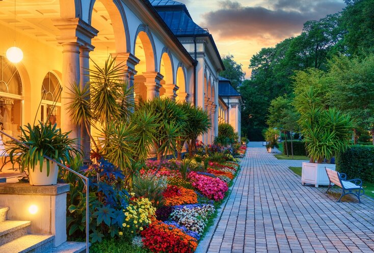 Sie sehen ein historisches Gebäude im Kurpark von Bad Steben am Abend mit Beleuchtung. Vor dem Gebäude sind bunte Blumen angepflanzt.