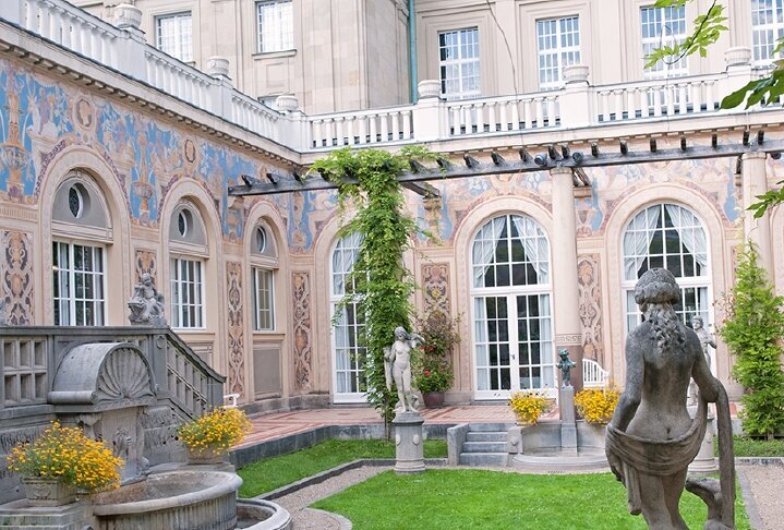 Sie sehen den Schmuckhof des Regentenbaus in Bad Kissingen. Im Vordergrund ist die Statue einer Frau von hinten und eine große Rasenfläche zu sehen und dahinter der Regentenbau.