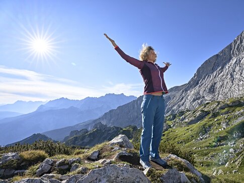Hier sehen Sie eine Frau mit blondem Haar und ausgestreckten Armen am Berg beim Atmen.
