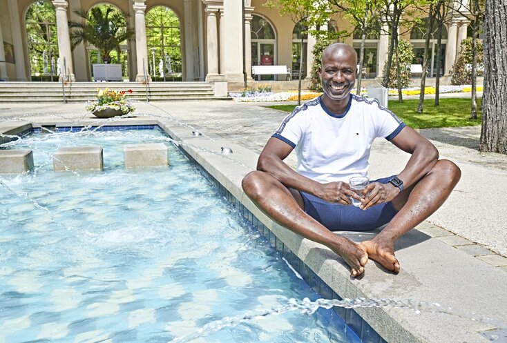 Hier sehen Sie Michel Frenzel-Assih, wie er neben einem Brunnen sitzt und ein Glas Wasser in der Hand hält