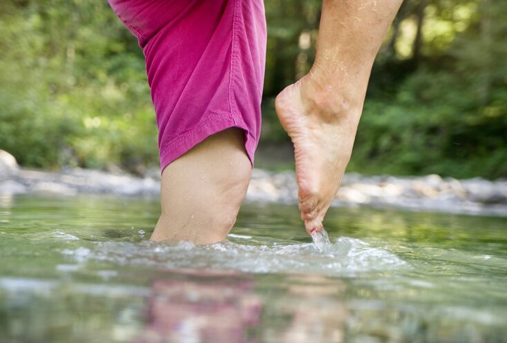 Hier sehen sie die Beine einer Frau, die im kniehohen Wasser steht. Sie kneippt im Storchenschritt. Im Hintergrund erkennt man, dass die Frau in der freien Natur ist. Es sind Bäume und Büsche zu sehen. 