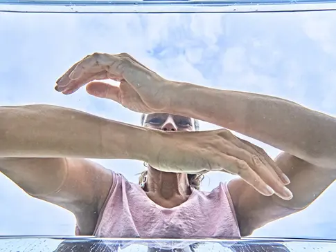 Hier sehen Sie eine Frau mit pinkem Shirt, welche ihre Arme in klares Wasser taucht. Die Aufnahme wurde aus dem Wasser, unterhalb der Frau, aufgenommen.