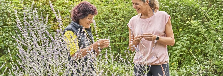 Hier sehen Sie zwei Frauen in einem Kräutergarten, die gerade Kräuter ernten.