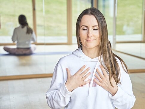 GESUNGES BAYERN-Expertin Susanne Lindlbauer sitzt mit einem weißen Pullover im Schneidersitz auf dem Holzfußboden und meditiert. Sie hat die Augen geschlossen, die langen braunen Haare fallen ihr über die linke Schulter und die Hände liegen auf Ihrer Brust.