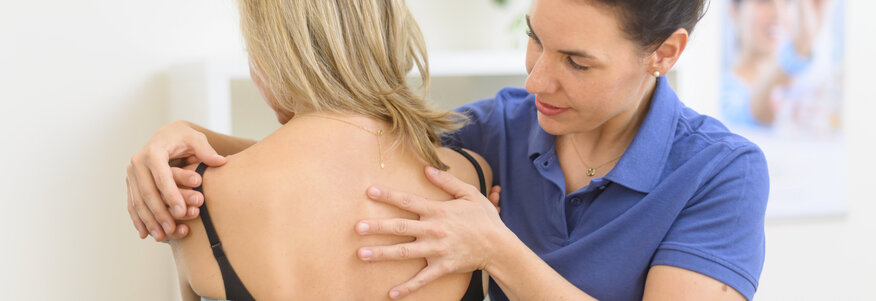 Hier sehen Sie eine Frau die mit dem Rücken zu uns sitzt und ein Handtuch umgewickelt hat. Eine Therapeutin steht neben ihr und behandelt den Rücken der Patientin. 