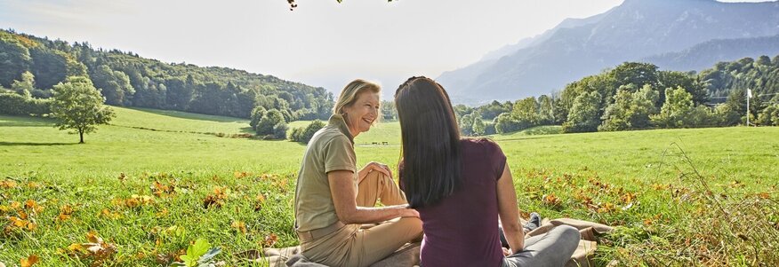 Hier sehen sie zwei Frauen auf einer Wiese sitzen. Die eine Frau erklärt der anderen etwas. Beide sind mit dem Rücken zu uns gedreht. Vor den zwei Frauen erstreckt sich ein Bergpanorama. . 