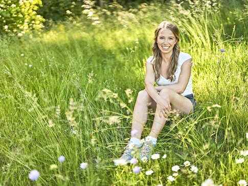 Sie sehen die GESUNDES BAYERN-Expertin Sarah Reinmold in einer Wiese sitzen und in die Kamera lächeln. Sie trägt ein weißes T-Shirt, eine kurze Hose und Turnschuhe.