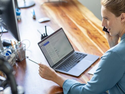 Eine Frau im Homeoffice sitzt an ihrem Schreibtisch vor ihrem Laptop und telefoniert.