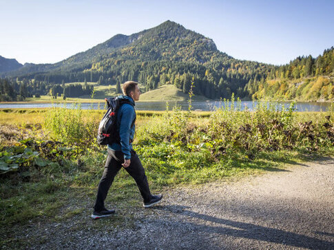 Hier sehen Sie den ehemaligen Profi-Fußballer Philipp Lahm. In Wanderbekleidung geht er auf einem Schotterweg spazieren. Hinter ihm sind Berge und ein See zu sehen. 