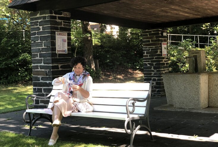 Sie sehen eine Frau, die auf einer Bank vor dem Trinkbrunnen im Kurpark in Bad Steben sitzt und sich ein Glas Wasser einschenkt.