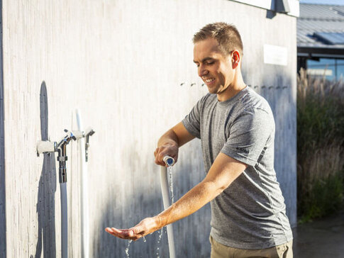 Hier sehen Sie den ehemaligen Profi-Fußballer Philipp Lahm. Er steht an einer Hauswand, aus der ein Wasserhahn steht. Er kühlt sich mit dem Wasserschlauch die Arme ab.