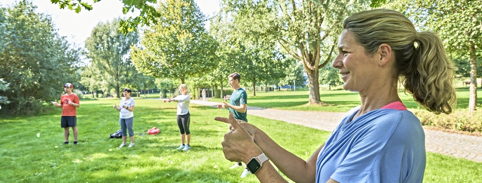 Sie sehen eine blonde Frau mit hellblauem T-Shirt im Vordergrund auf einer Wiese stehen, die mit beiden Händen eine Fingerübung macht. Sie macht mit beiden Händen gleichzeitig unterschiedliche Bewegungen. Im Hintergrund sind noch vier weitere Personen zu sehen, die ebenfalls an der Übung teilnehmen und bunte Sportkleidung tragen. Es sind ebenfalls hinter den Menschen Bäume und Wege zu sehen.