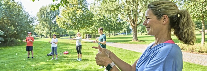 Sie sehen eine blonde Frau mit hellblauem T-Shirt im Vordergrund auf einer Wiese stehen, die mit beiden Händen eine Fingerübung macht. Sie macht mit beiden Händen gleichzeitig unterschiedliche Bewegungen. Im Hintergrund sind noch vier weitere Personen zu sehen, die ebenfalls an der Übung teilnehmen und bunte Sportkleidung tragen. Es sind ebenfalls hinter den Menschen Bäume und Wege zu sehen.