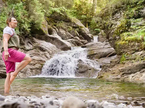 Hier sehen Sie einen kleinen Bergfluss. Bayern Botschafterin Christine Waibel-Beer steht an einer seichten Stelle im Wasser und kneippt im Storchenschritt. 