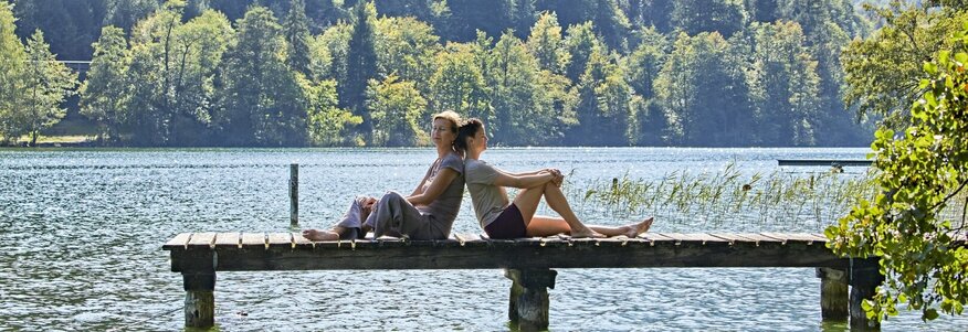 Hier sehen Sie einen Steg von der Seite der in einen großen See läuft. Auf dem Steg sitzen eine Frau und ein Mann, Rücken an Rücken und genießen das schöne Wetter. Hinter ihnen sind Berge und Wald zu sehen. 