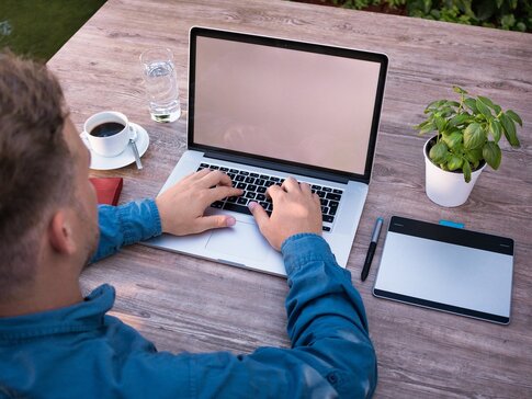 Hier sehen Sie einen Mann schräg von hinten, welcher draußen an einem Gartentisch an seinem Laptop sitzt und arbeitet. Neben ihm hat er noch ein Taplat sowie Stifte liegen. Auch einen Kaffe, ein Glas Wasser und eine Topfpflanze steht neben ihm auf dem Tisch. 