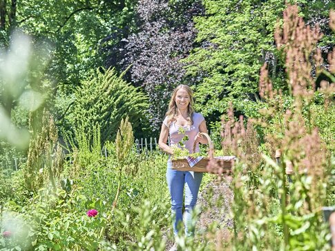 Hier sehen Sie eine junge Frau, die mit einem Korb voller Kräuter durch einen Kräutergarten spaziert.