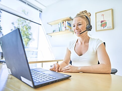 Sie sehen die GESUNDES BAYERN-Expertin Sarah Reinmold in ihrem Büro am Schreibtisch sitzen. Vor ihr steht ein geöffneter Laptop auf der Holztischplatte des Schreibtisches und sie trägt ein weißes T-Shirt und ein Headset. Im Hintergrund sind Bilder an der weißen Wand und eine große Fensterfront zu sehen.