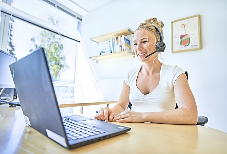 Sie sehen die GESUNDES BAYERN-Expertin Sarah Reinmold in ihrem Büro am Schreibtisch sitzen. Vor ihr steht ein geöffneter Laptop auf der Holztischplatte des Schreibtisches und sie trägt ein weißes T-Shirt und ein Headset. Im Hintergrund sind Bilder an der weißen Wand und eine große Fensterfront zu sehen.