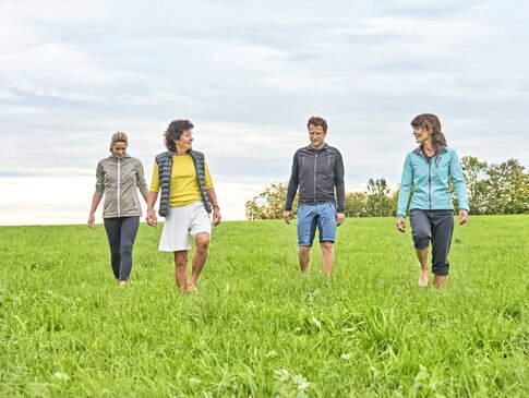 Eine Gruppe von drei Frauen und einem Mann wandern Barfuß durch eine feuchte Wiese.