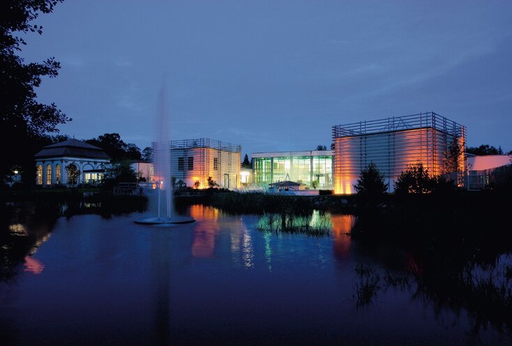 Sie sehen die Gebäude der Therme in Bad Steben am Abend in bunten Farben beleuchtet. Im Vordergrund ist ein See mit einer Wasserfontäne zu sehen.