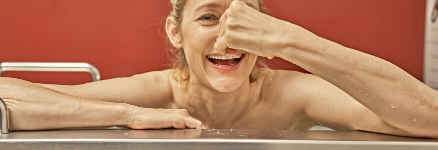 Hier sehen Sie eine Frau, die in einer Badewanne sitzt und sich lachend die Nase zuhält.