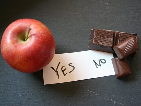 Auf der linken Bildhälfte sieht man einen Apfel. Rechts ein Stück Schokolade. Zwischen drin liegt ein Zettel mit dem Schriftzug Yes No.