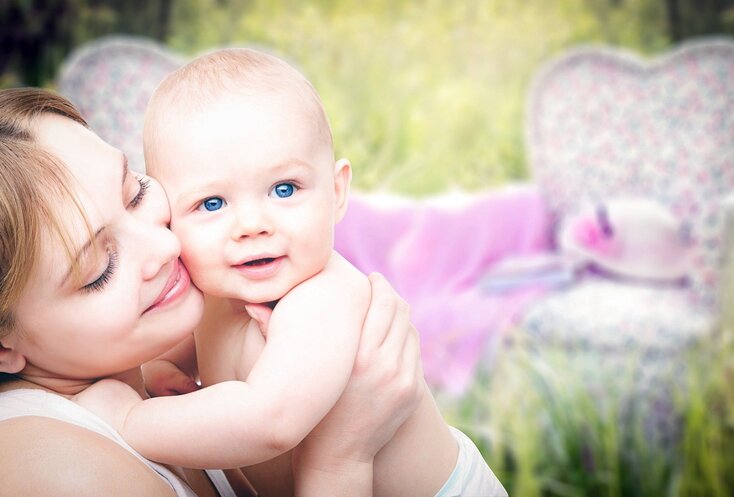 Hier sehen Sie eine Mutter mit einem kleinen Kind im Arm. Sie sitzen auf einer Wiese. Hinter ihnen sind undschwarf weiter Sitzmöglichkeiten zu sehen. 