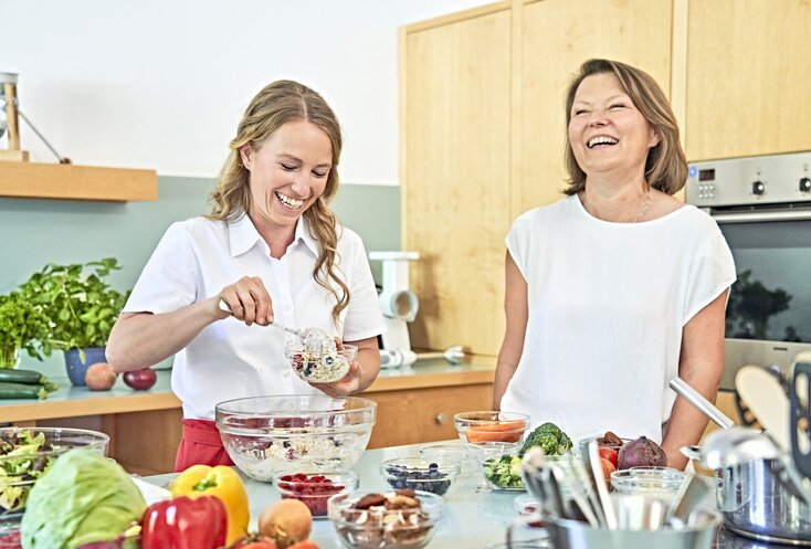 Sie sehen links Ernährungsberaterin Sarah Reinmold zusammen mit einer anderen Frau in der Lehrküche der Malteser Klinik von Weckbecker beim Kochen. Beide Frauen tragen weiße Oberteile und lachen. Im Vordergrund ist verschiedenes Gemüse und Obst, sowie Kochgeschirr zu sehen. Im Hintergrund sind helle holzfarbene Küchenfronten und der Backofen zu sehen.