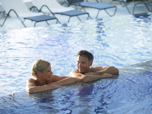 Hier sehen Sie ein Paar in einem Pool. Beide halten sich am Rand des Pools mit den Oberarmen fest. Sie unterhalten sich. Im Hintergrund sind Liegen zu sehen. 