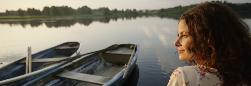 Hier sehen Sie eine Frau die auf einem Steg sitzt. Neben ihr im Wasser sind kleinere Padelbote. Sie sitzt mit dem Rücken zu uns und hat ihren Kopf leicht zu uns gedreht. Die Atmosphäre deutet auf einen Sonnenuntergang im Sommer hin. 