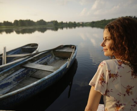 Hier sehen Sie eine Frau die auf einem Steg sitzt. Neben ihr im Wasser sind kleinere Padelbote. Sie sitzt mit dem Rücken zu uns und hat ihren Kopf leicht zu uns gedreht. Die Atmosphäre deutet auf einen Sonnenuntergang im Sommer hin. 