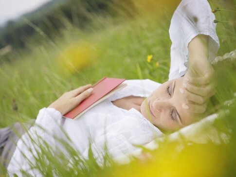 Hier sehen Sie eine Frau, die mit geschlossenen Augen im hohen Gras liegt und ein Buch auf dem Bauch liegen hat.