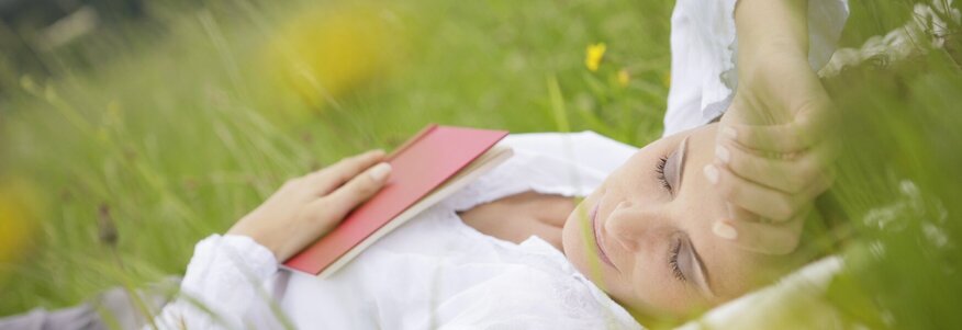 Hier sehen Sie eine Frau, die mit geschlossenen Augen im hohen Gras liegt und ein Buch auf dem Bauch liegen hat.