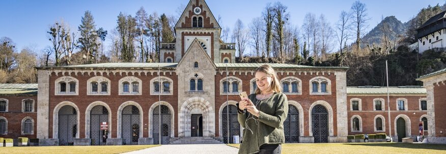 Im Hintergrund ist ein großes Gebäude aus rotem Backstein zu sehen. Das ist die Alte Saline. Hier wird seit mehr als 150 Jahren salzhaltige AlpenSole aus den Bergen bei Bad Reichenhall gefördert um es für Kur- und Gesundheitszwecke zu nutzen. Auf dem Platz vor dem Gebäude ist eine Frau zu sehen, die die Saline besucht hat.