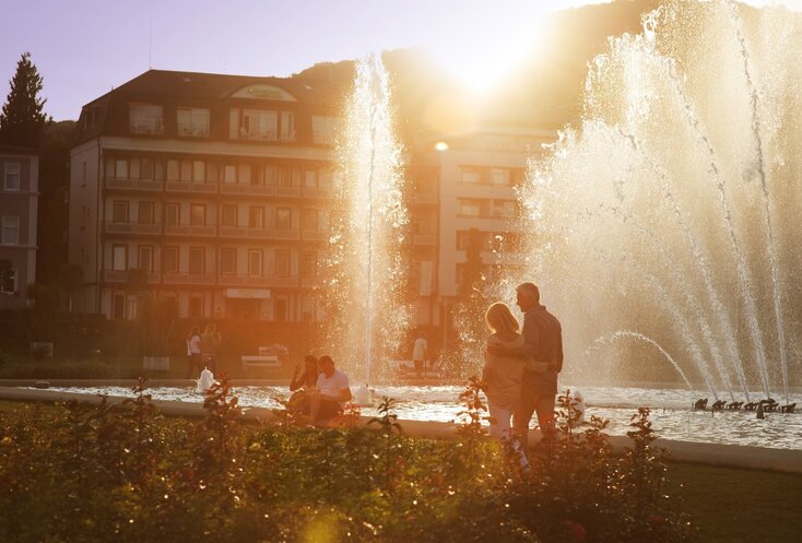 Auf der rechten Seite ist eine Wasserfontäne im Rosengarten in Bad Kissingen zu sehen, dahinter ein großes Gebäude hinter dem die Sonne untergeht.