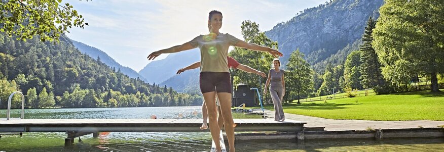 Hier sehen Sie Personen über einen Holzbalken balancieren. Der Holzalken ist über einer kleinen Wasserstelle. Im Hintergrund sind Berge und Wälder zu sehen. 