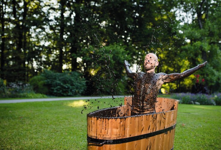 Hier sehen Sie einen Mann in einer Moorbadewanne. Die Wanne steht auf einer Wiese im Freien. Der Mann lacht und wirft das Moor in die Luft.