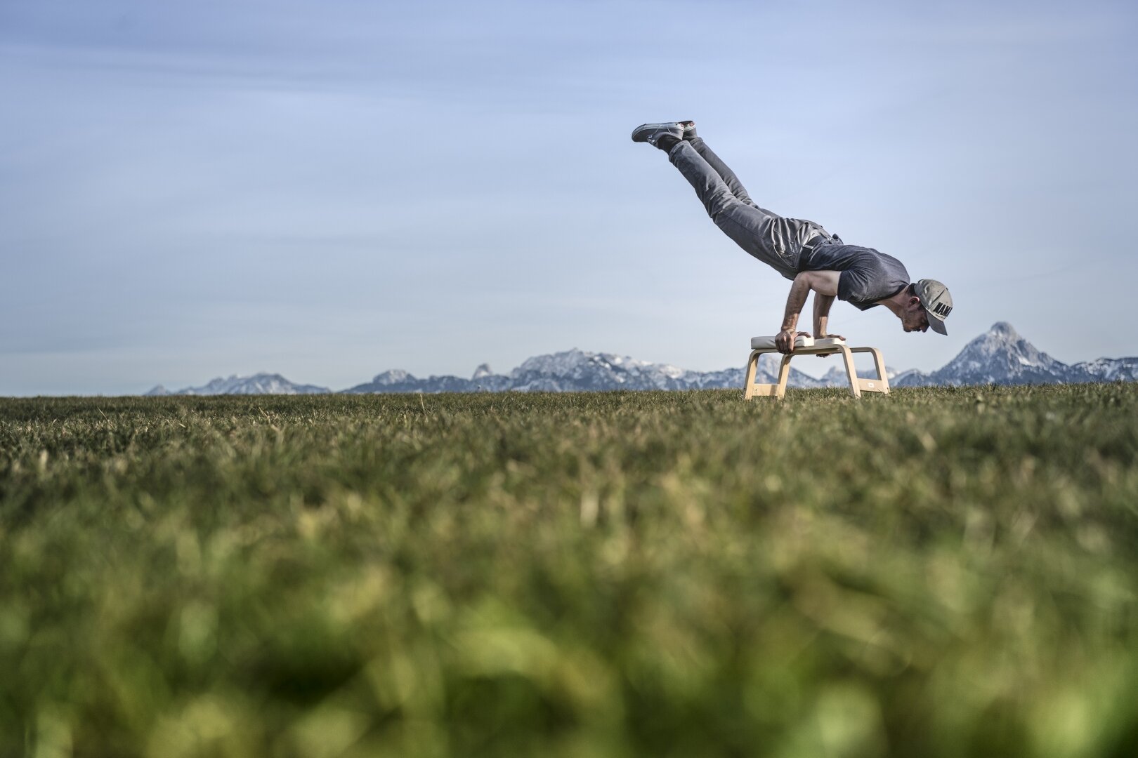 Hier sehen Sie den Bayern Botschafter Kilian Trenkle. Auf einer Wiese macht er Yogaübungen mit einem Yogahocker. Hinter ihm ist eine klassische bayerische Landschaft zu sehen.