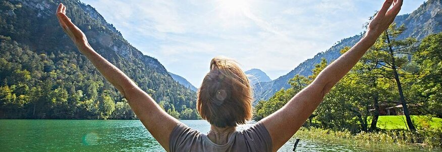 Hier sehen Sie eine Frau, welche vor einem See steht. Sie hat ihre Arme über die Seite nach oben. 