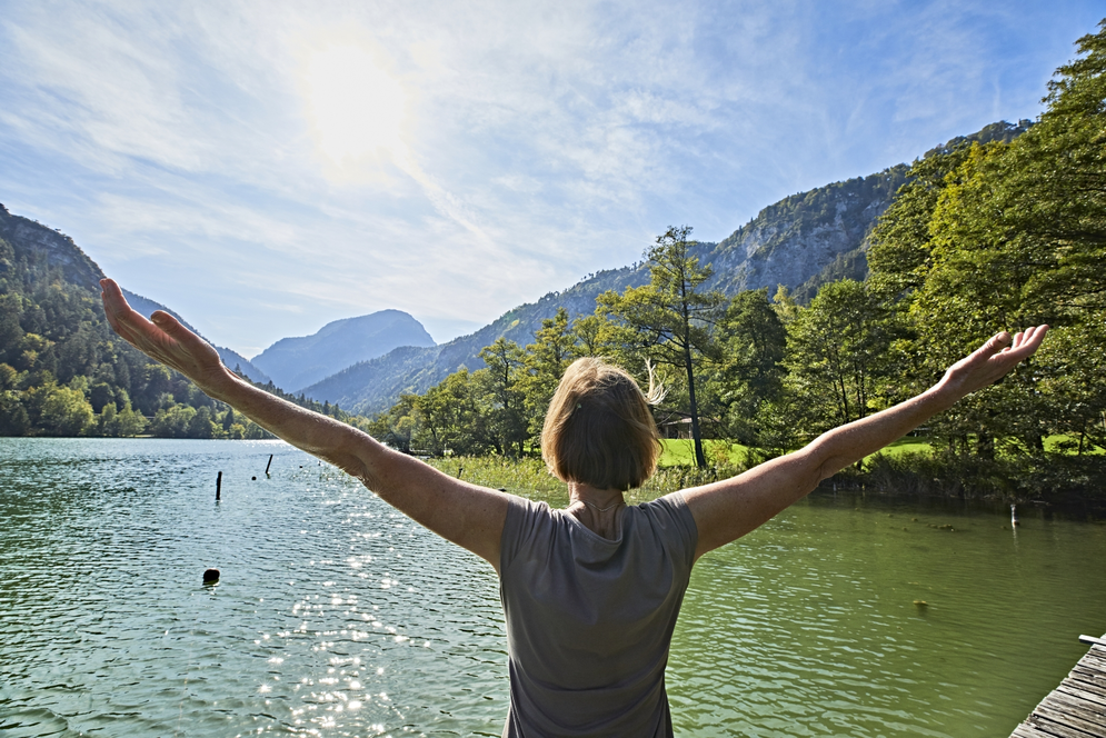 Steg, Natur, Energie Hier sehen Sie eine Frau die mit dem Rücken zu uns am Seeufer steht und beide Arme über die Seite nach oben hebt. Die Frau blickt auf den See und auf die dahinterliegenen Berge und Wälder.