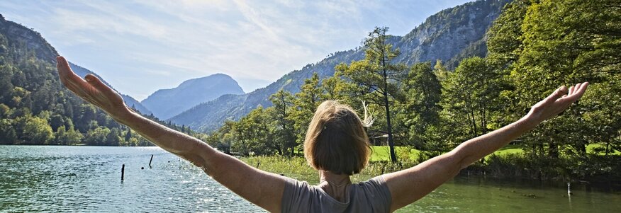 Hier sehen Sie eine Frau die mit dem Rücken zu uns am Seeufer steht und beide Arme über die Seite nach oben hebt. Die Frau blickt auf den See und auf die dahinterliegenen Berge und Wälder. 