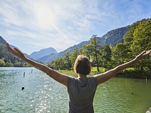 Hier sehen Sie eine Frau die mit dem Rücken zu uns am Seeufer steht und beide Arme über die Seite nach oben hebt. Die Frau blickt auf den See und auf die dahinterliegenen Berge und Wälder. 