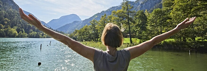 Hier sehen Sie eine Frau die mit dem Rücken zu uns am Seeufer steht und beide Arme über die Seite nach oben hebt. Die Frau blickt auf den See und auf die dahinterliegenen Berge und Wälder. 