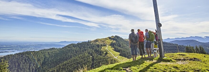 Hier sehen Sie eine Gruppe an Wanderern, die das Gipfelkreuz erreicht haben und auf eine Berglandschaft blicken. 