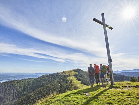 Hier sehen Sie eine Gruppe an Wanderern, die das Gipfelkreuz erreicht haben und auf eine Berglandschaft blicken. 