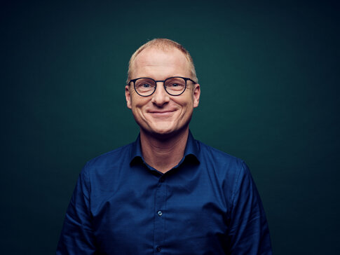 Hier sehen Sie ein Portraitfoto von dem #gesundkannjeder Experten Dr. Manfred Wagner. Er trägt ein blaues Hemd und ein Brille. Der Hintergrund ist einfarbig und dunkel. 